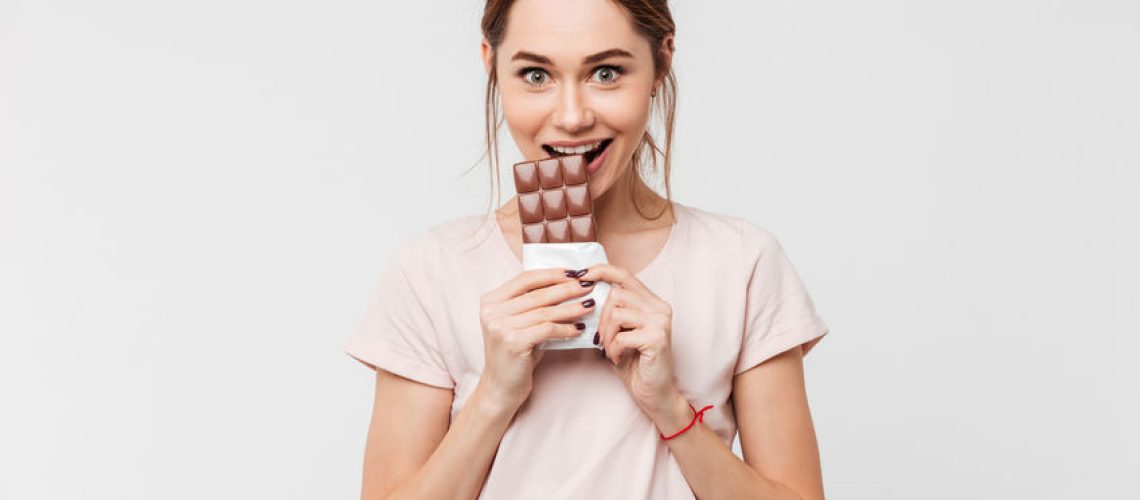 Portrait of a smiling pretty girl eating chocolate bar and looking at camera isolated over white background