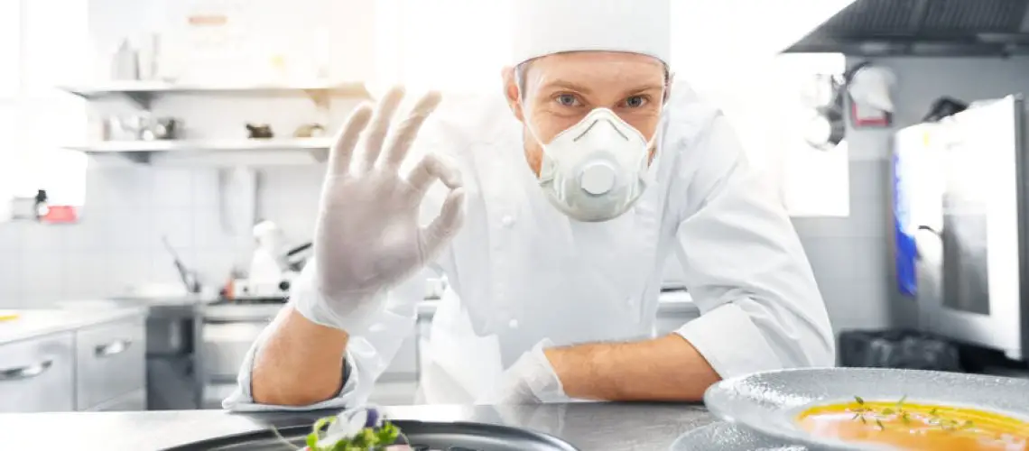 health, safety and pandemic concept - male chef cook wearing face protective mask or respirator for protection from virus disease with plate of soup and salad showing ok sign at restaurant kitchen